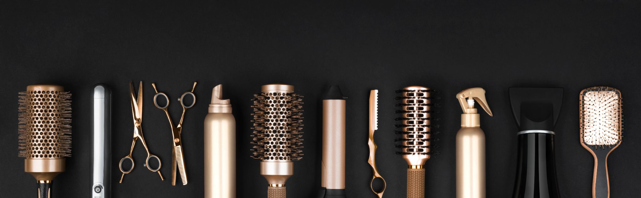 Professional hair styling tools arranged in a row including brushes, scissors, sprays, and combs on black background