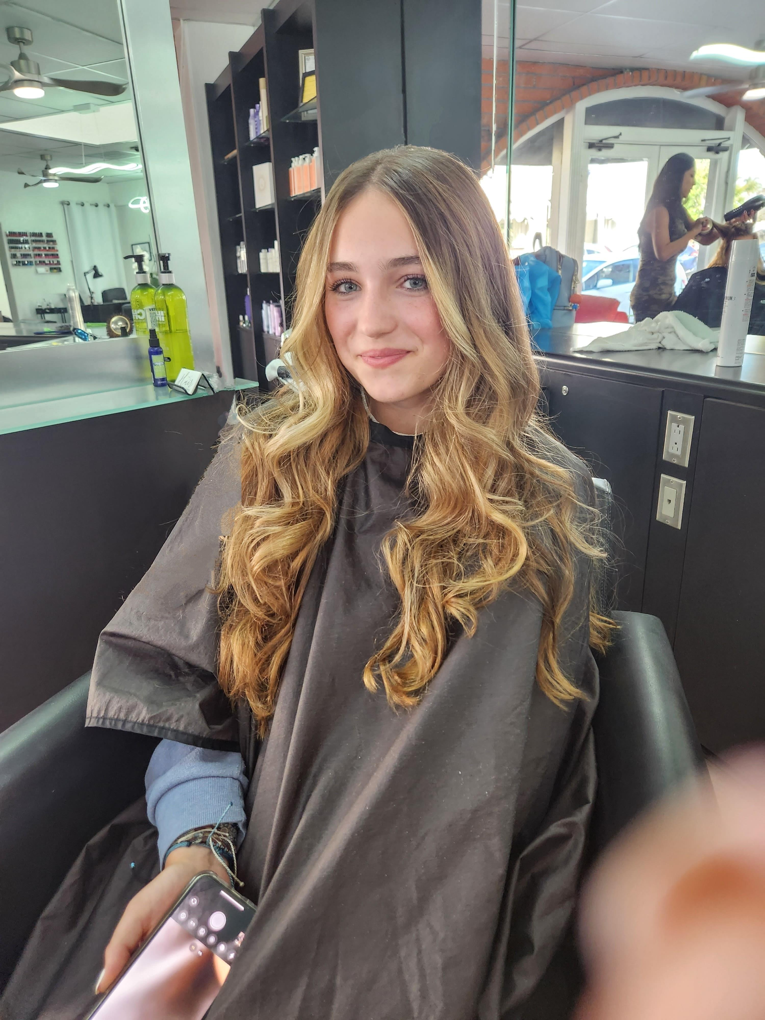Woman with long wavy brunette hair smiling in a salon chair wearing a black cape