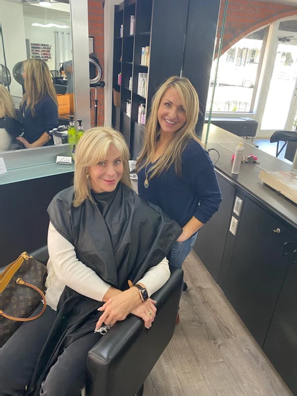 Two women smiling at a salon station with mirror and styling products visible